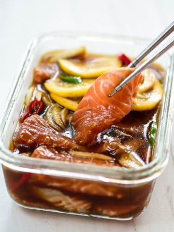 Slices salmon in a soy sauce marinade in a rectangle glass container a slice being picked up with chopsticks