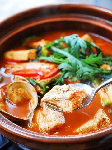 Korean spicy fish stew with clams, tofu and green veggies in an earthenware with a fish piece in a spoon